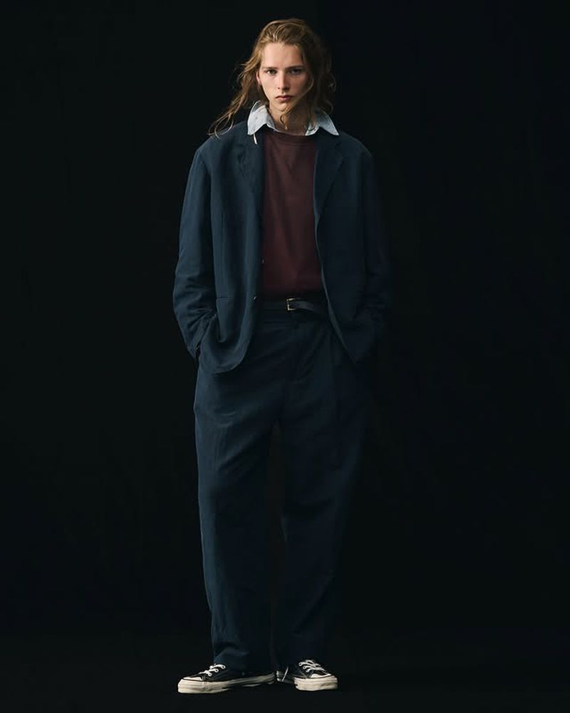Full-length shot of an adult male fashion model posing in a navy blue suit, maroon tee shirt, and black sneakers against a black backdrop.