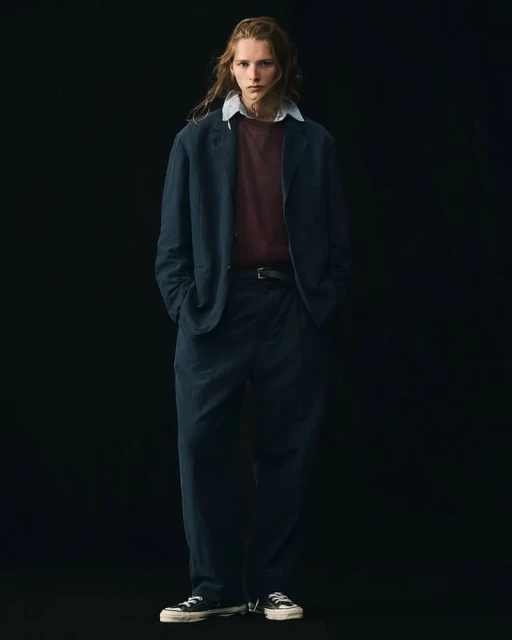 Full-length shot of an adult male fashion model posing in a navy blue suit, maroon tee shirt, and black sneakers against a black backdrop.