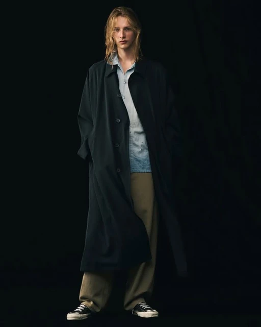 A fair-skinned adult male with long blonde hair poses in a black trench coat, button-up shirt, and trousers against a dark background.