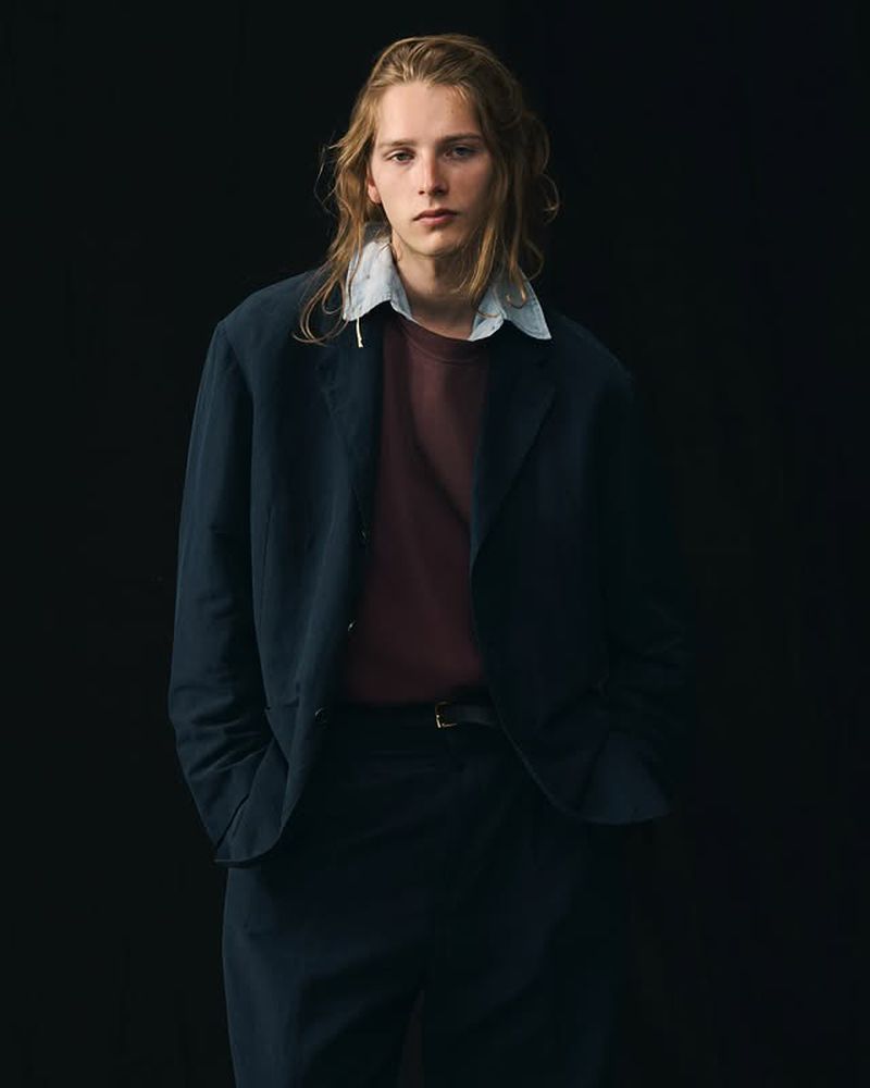 Portrait of a young adult male with long blond hair wearing a navy jacket, burgundy t-shirt, and dress pants against a dark background.