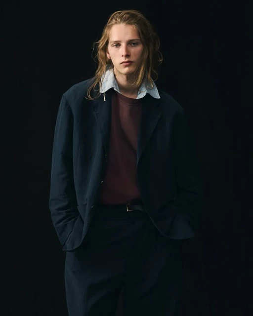 Portrait of a young adult male with long blond hair wearing a navy jacket, burgundy t-shirt, and dress pants against a dark background.