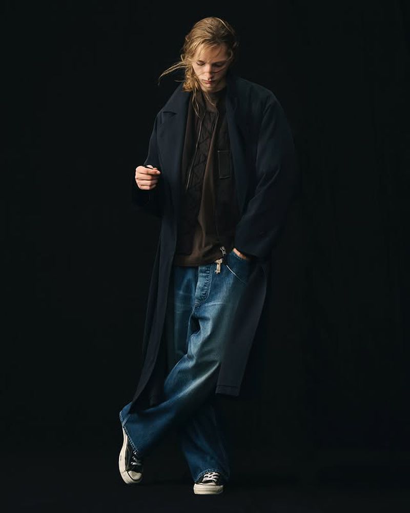 Model wearing a dark long coat, brown shirt, and wide-leg denim jeans against a solid black background.