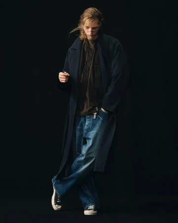 Model wearing a dark long coat, brown shirt, and wide-leg denim jeans against a solid black background.