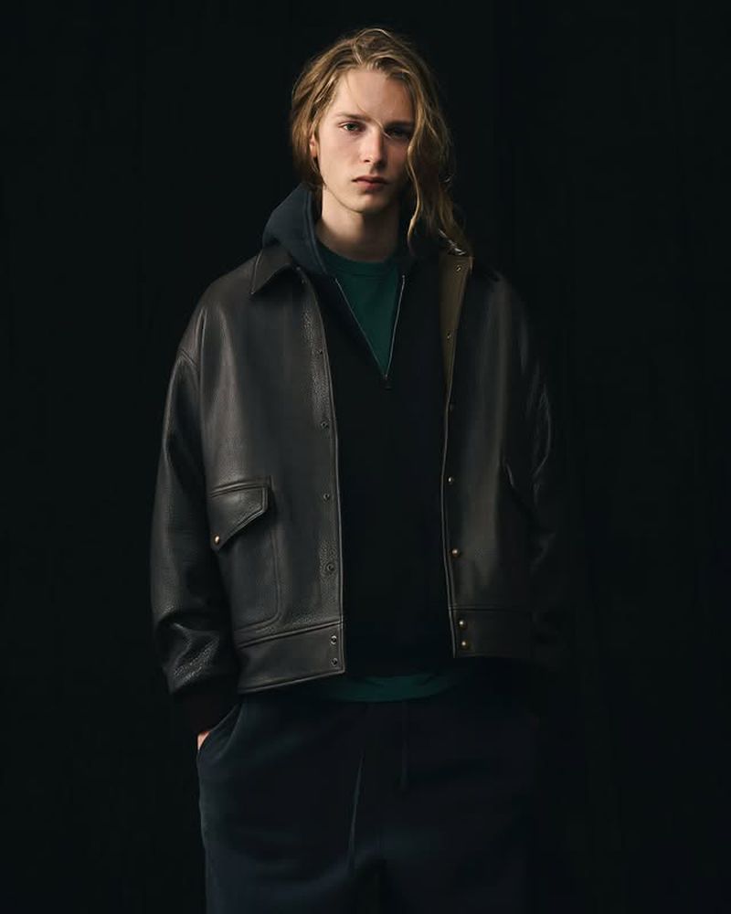 Young man with long blond hair poses in a dark leather jacket, dark hooded top, and pants against a black backdrop in a studio shot.