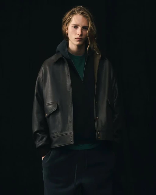 Young man with long blond hair poses in a dark leather jacket, dark hooded top, and pants against a black backdrop in a studio shot.