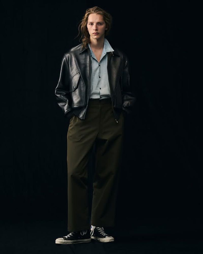 Young adult poses in black leather jacket and olive slacks in a studio, showcasing a blend of modern fashion and classic style.