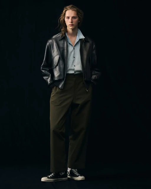 Young adult poses in black leather jacket and olive slacks in a studio, showcasing a blend of modern fashion and classic style.