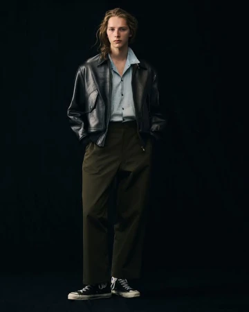 Young adult poses in black leather jacket and olive slacks in a studio, showcasing a blend of modern fashion and classic style.
