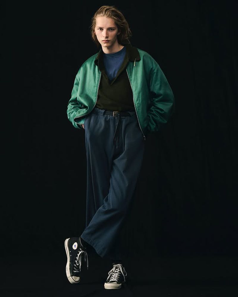 Young adult man in green jacket, blue shirt and pants, and black sneakers posing in a dimly lit photo studio against a black backdrop.