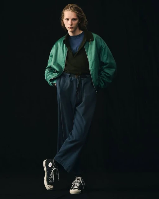 Young adult man in green jacket, blue shirt and pants, and black sneakers posing in a dimly lit photo studio against a black backdrop.