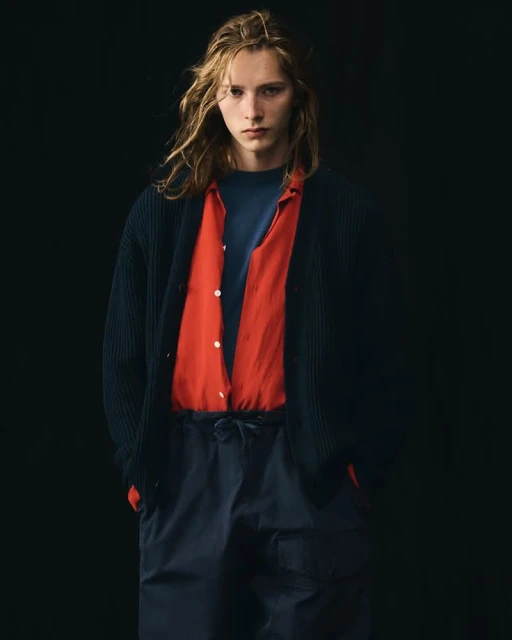 Blond male fashion model in a blue cardigan, red shirt, and blue drawstring pants poses against a dark background in a portrait-style studio shot.