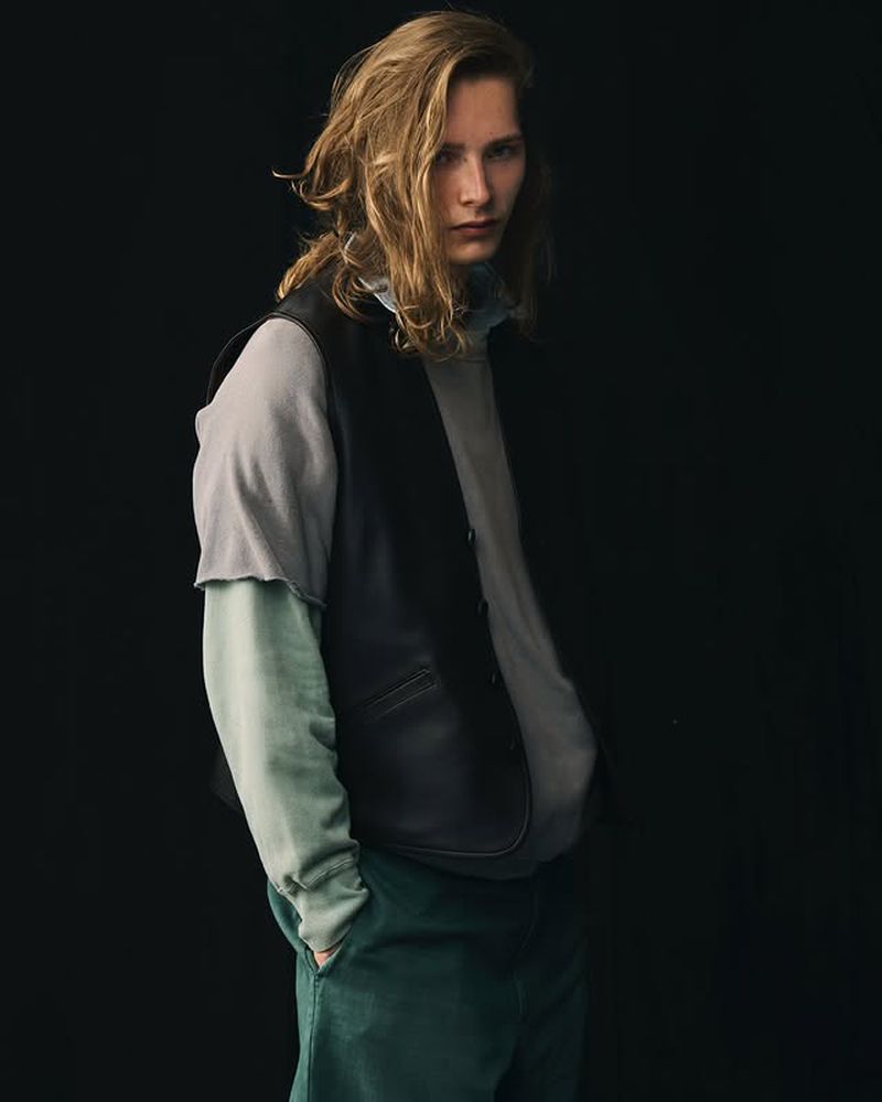 Blond male model in a layered grey and blue outfit with a black vest, standing in front of a dark background, looking at the camera.