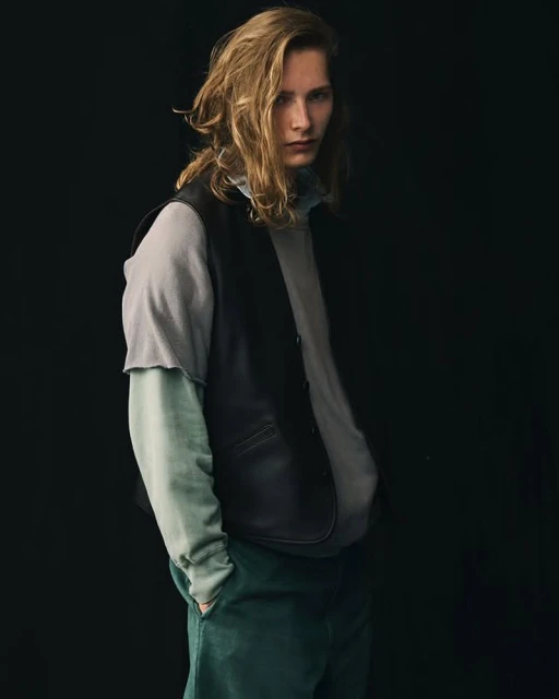 Blond male model in a layered grey and blue outfit with a black vest, standing in front of a dark background, looking at the camera.