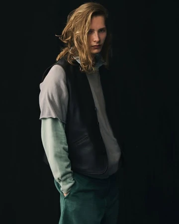 Blond male model in a layered grey and blue outfit with a black vest, standing in front of a dark background, looking at the camera.