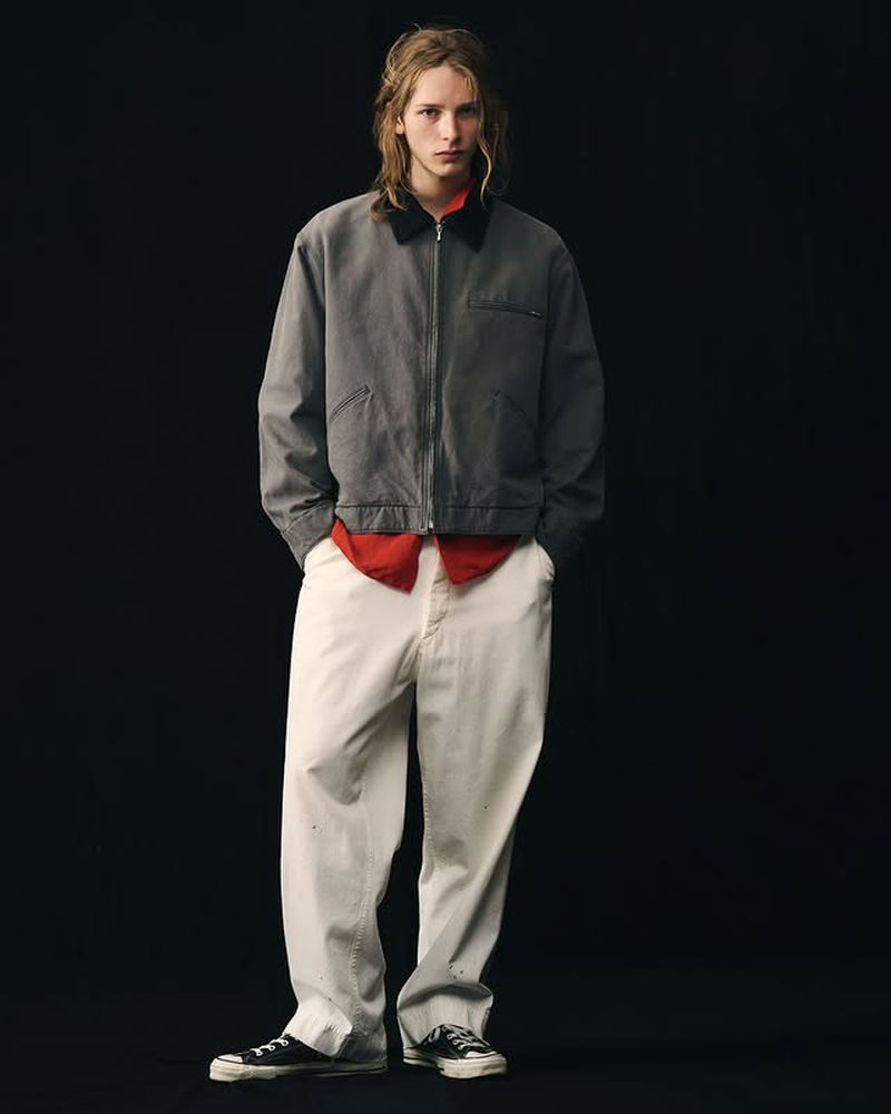 Full shot of a young adult male model in a grey jacket, red collared shirt, and white pants in a minimalist fashion pose on black.