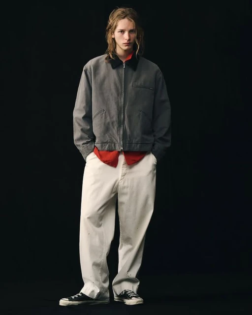 Full shot of a young adult male model in a grey jacket, red collared shirt, and white pants in a minimalist fashion pose on black.