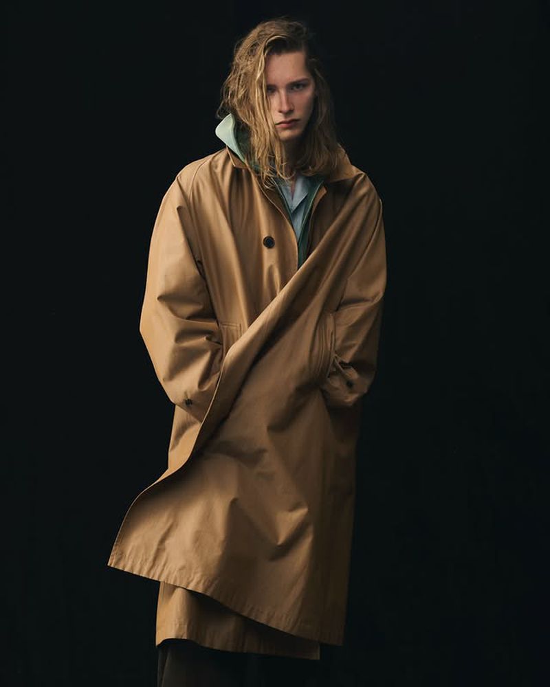 Studio shot of a blond adult male wearing a tan trench coat over a light blue shirt and hoodie, standing against a black background.