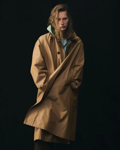 Studio shot of a blond adult male wearing a tan trench coat over a light blue shirt and hoodie, standing against a black background.