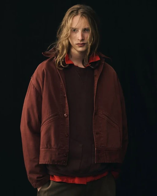 Portrait of a young adult male with long blond hair wearing a rust-colored jacket, brown sweater, and green pants in a dark studio.