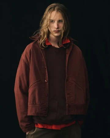 Portrait of a young adult male with long blond hair wearing a rust-colored jacket, brown sweater, and green pants in a dark studio.