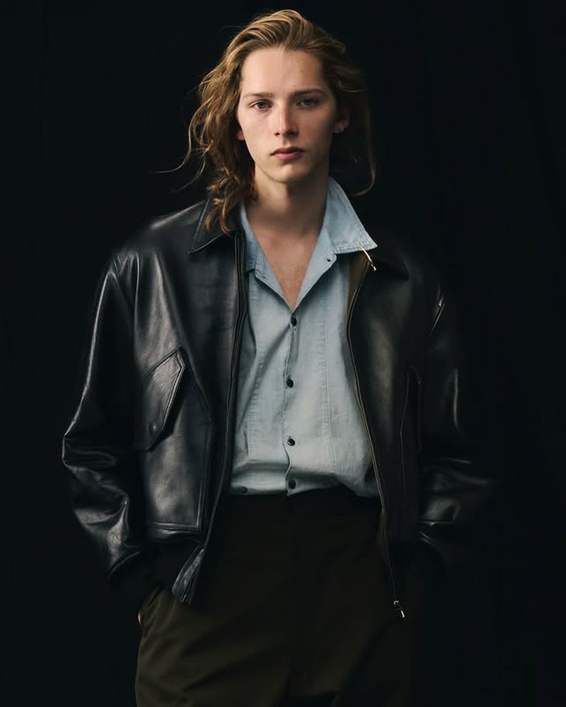 Man with long blond hair wearing a black leather jacket and light blue shirt poses against a dark backdrop in a studio shot.