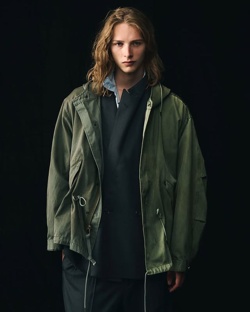 A long-haired male model poses in front of a dark background, wearing an olive green hooded jacket and dark grey top in a studio shot.