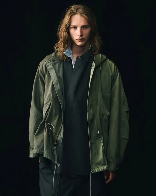A long-haired male model poses in front of a dark background, wearing an olive green hooded jacket and dark grey top in a studio shot.