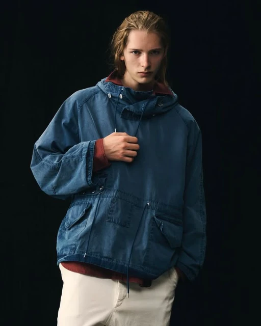 A fair-skinned young man with long blond hair models a blue hooded jacket and cream pants against a dark background in a studio shot.