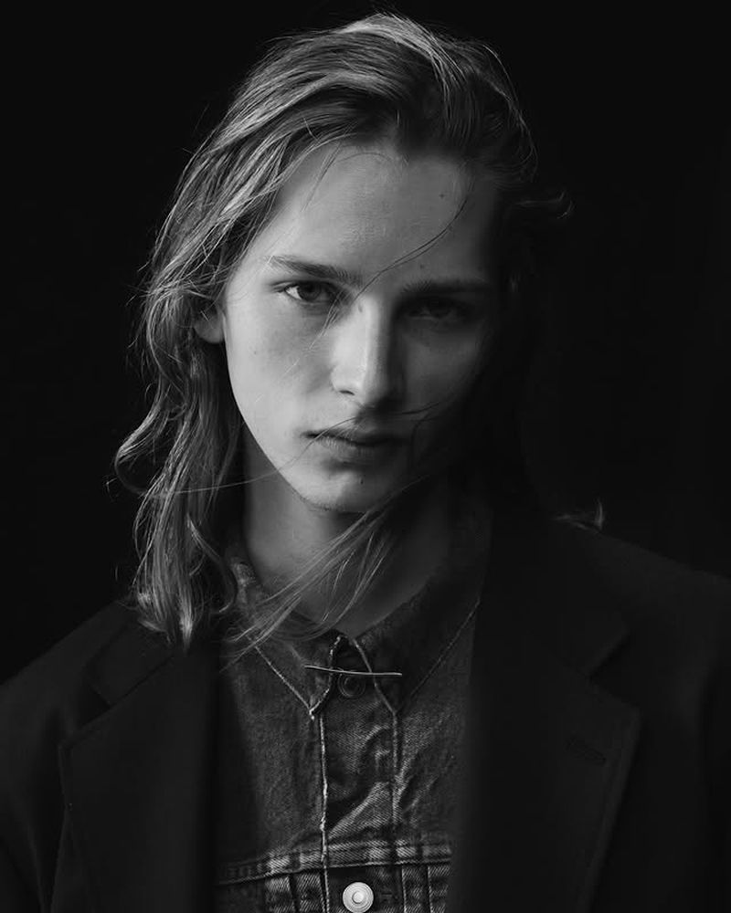 Portrait of an adult with long hair, wearing a denim shirt and blazer, posing against a black backdrop with diffused studio lighting.