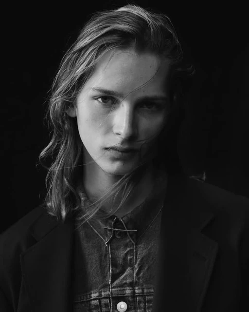Portrait of an adult with long hair, wearing a denim shirt and blazer, posing against a black backdrop with diffused studio lighting.