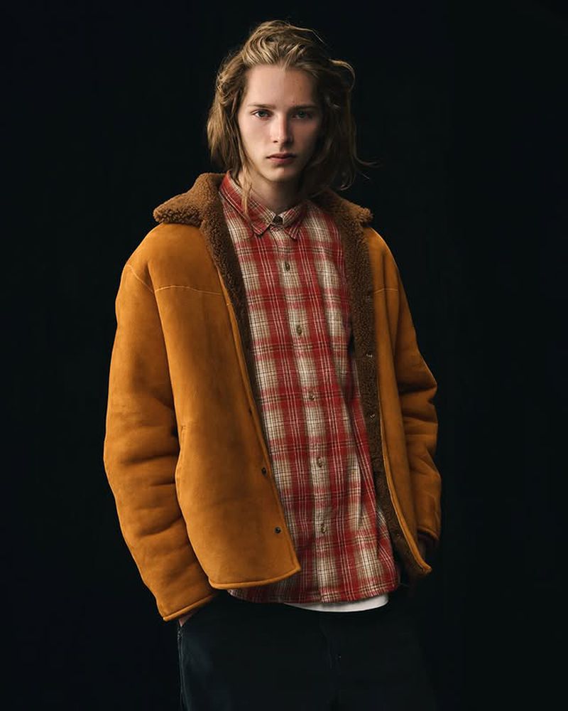 Adult male with medium-length blond hair wearing a tan jacket with fur collar and a red plaid shirt against a dark background.