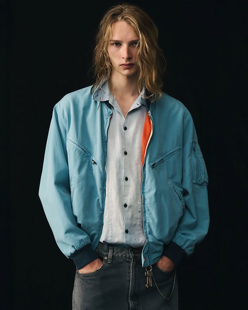 Fashion portrait of a young adult man with long blonde hair wearing a light blue bomber jacket, light button-down shirt, and dark pants.