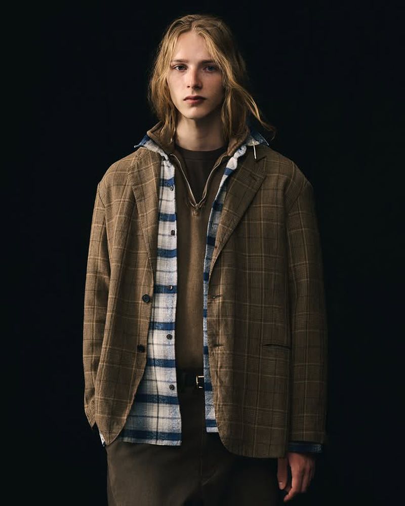 Young adult male in a brown plaid blazer, brown sweater, and blue and white plaid shirt, posing against a dark background in a studio shot.