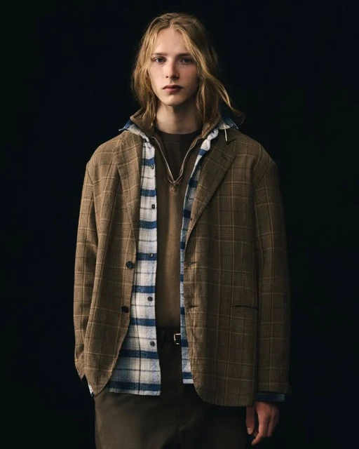 Young adult male in a brown plaid blazer, brown sweater, and blue and white plaid shirt, posing against a dark background in a studio shot.