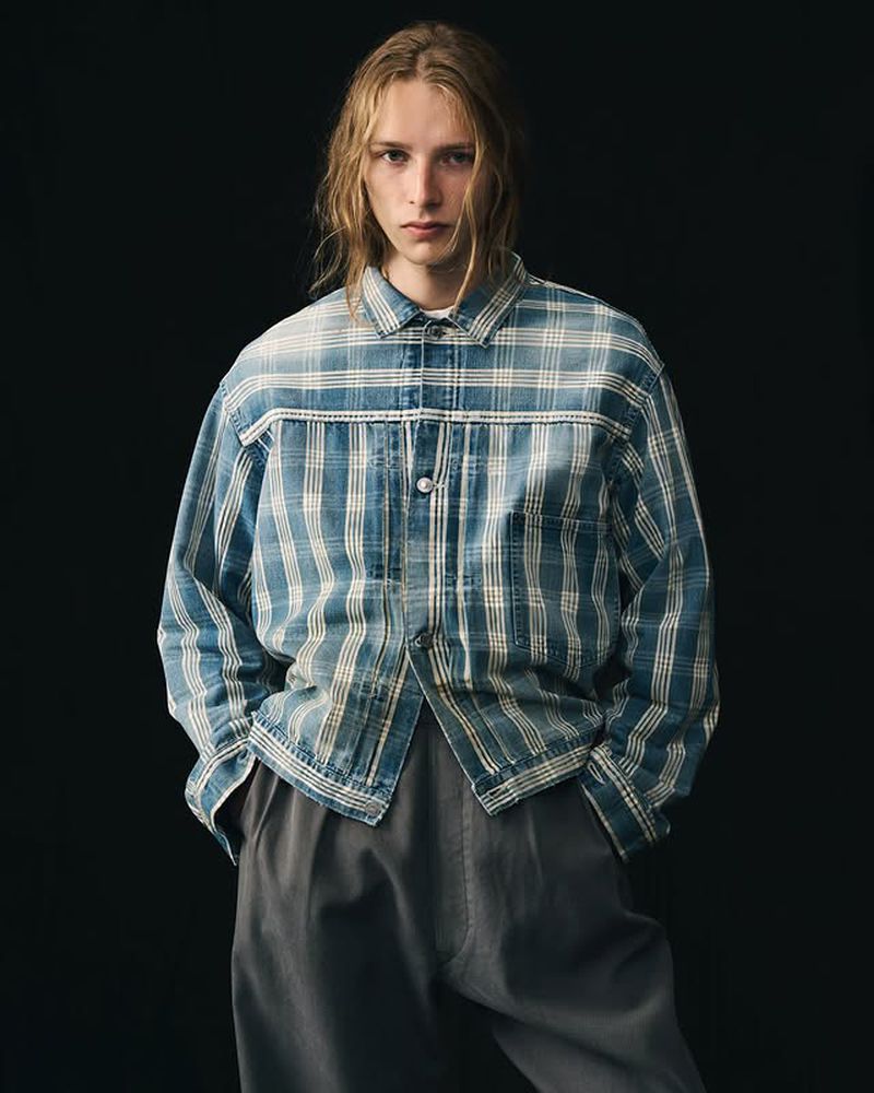 Blond-haired man in a blue and white plaid button-up shirt and grey trousers standing against a dark grey background in a studio portrait.