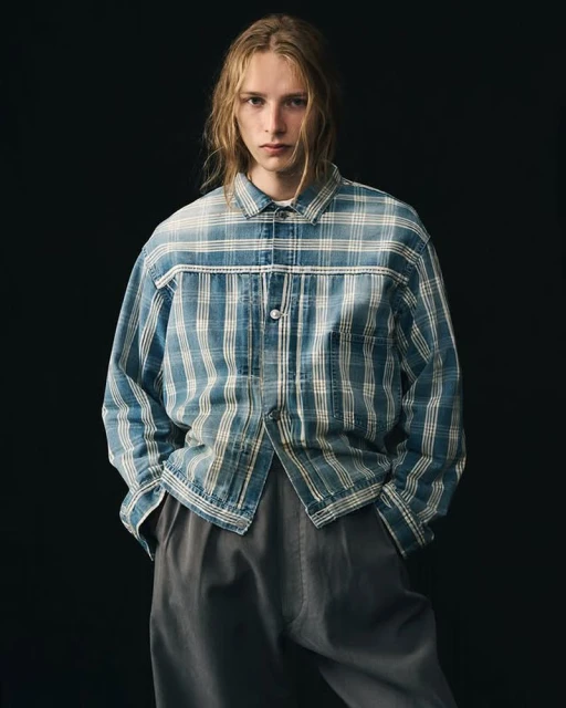 Blond-haired man in a blue and white plaid button-up shirt and grey trousers standing against a dark grey background in a studio portrait.