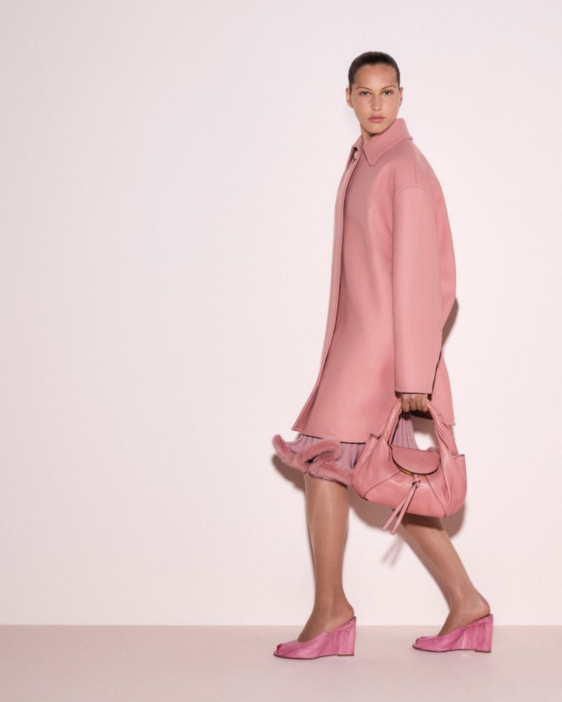 Woman in pink coat and dress carrying pink purse. Fashion photography in a studio setting against a pale pink background.