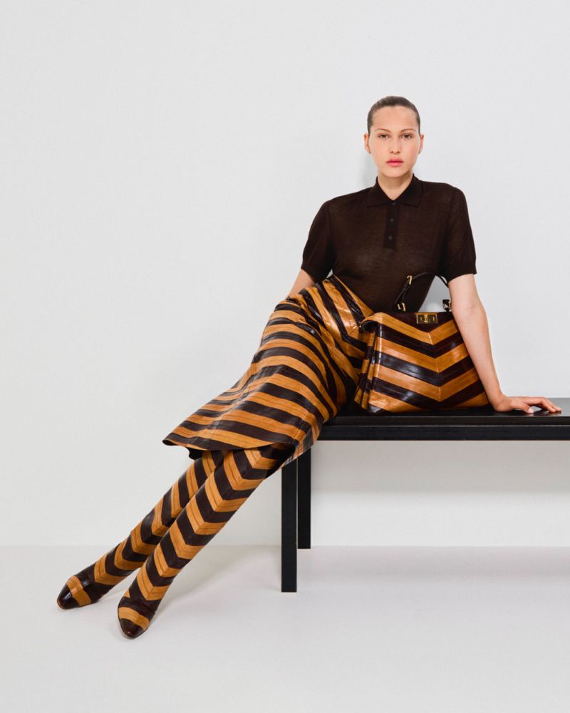 Woman poses in brown and black striped outfit, matching bag, and low-heeled shoes, sitting on a black bench in a bright photo studio.