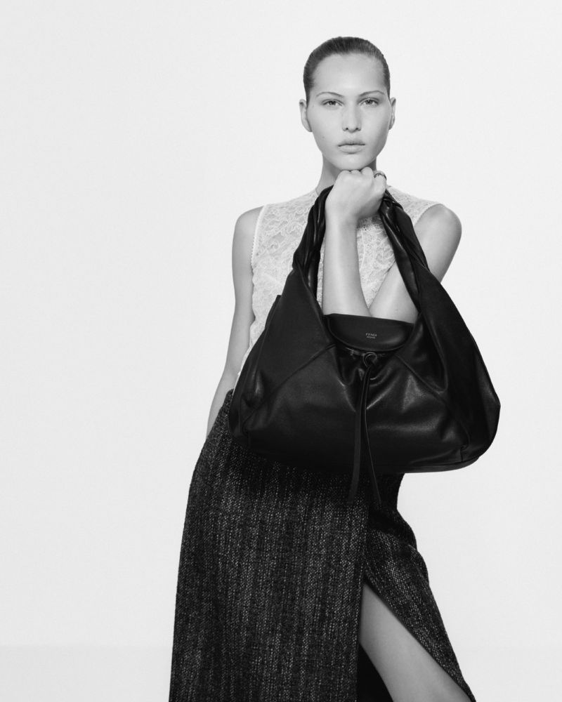 Model holding a large black leather handbag wearing a patterned skirt against a plain white background