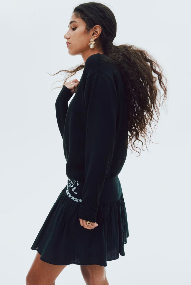 Fashion portrait of a young woman in black sweater and skirt, posing in a light studio with a side view showcasing her jewelry.