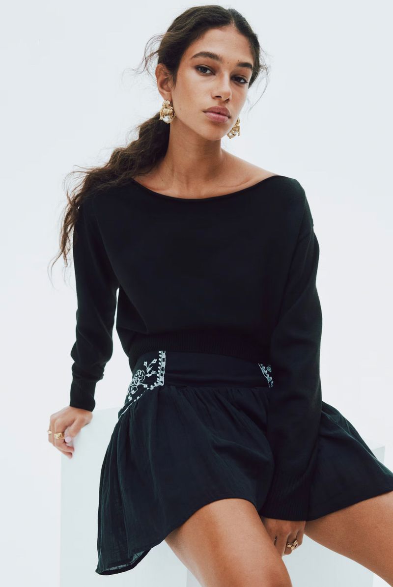 Brunette woman poses indoors wearing a black, long-sleeved top and skirt with decorative details, sitting on a white cube. Fashion shot.