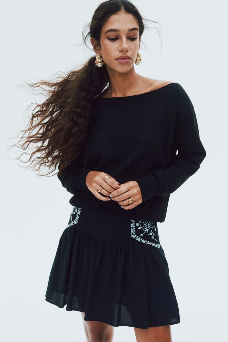 A woman is in a studio setting wearing a black sweater and a short black skirt with embroidery posing for a fashion shot.