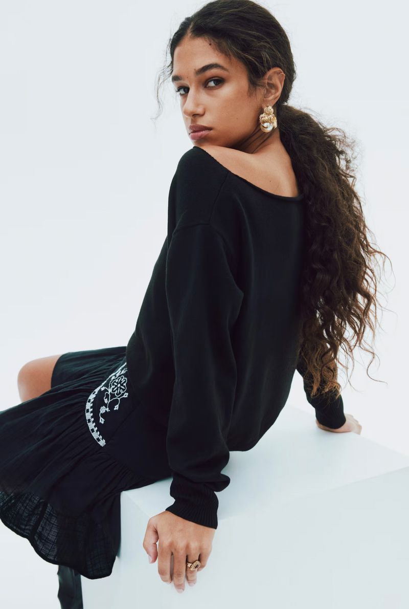 Portrait of a curly-haired, tan-skinned young woman in black sweater dress and gold earrings, looking over her shoulder in a studio.