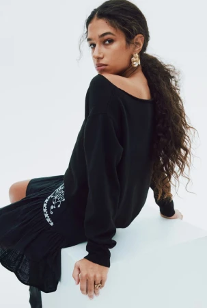Portrait of a curly-haired, tan-skinned young woman in black sweater dress and gold earrings, looking over her shoulder in a studio.