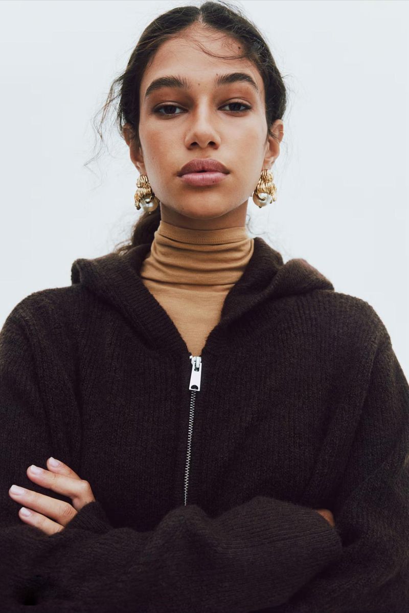 Woman wearing a brown knit hooded sweater and statement earrings poses against a white backdrop in a studio setting.