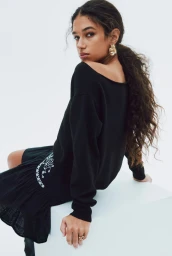 Tamar David seated on a white cube wearing an off-the-shoulder black sweater and ruffled skirt against a white studio background.