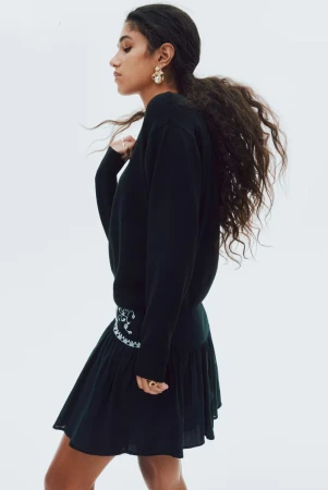 Model with long dark curly hair wearing a black long-sleeve top and skirt against a seamless white background