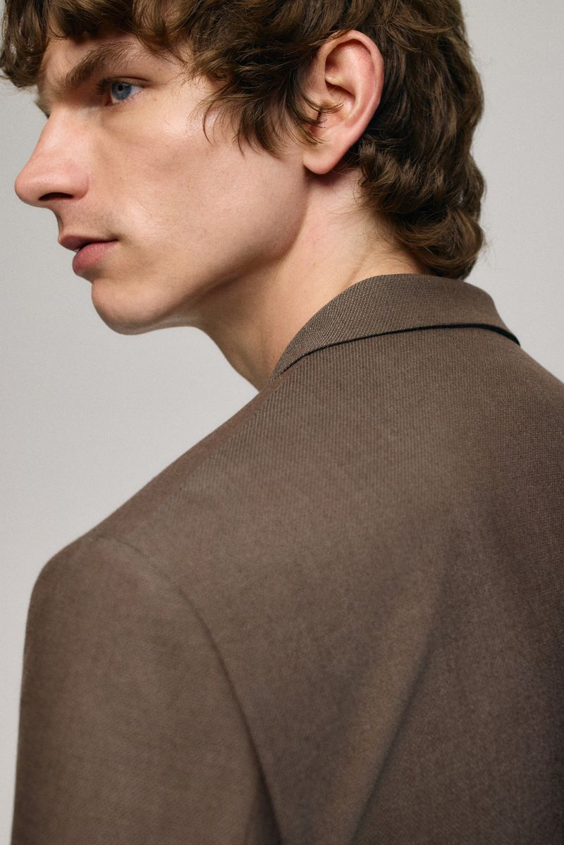 Close-up studio shot of a male model with wavy brown hair, wearing a brown blazer, gazing to the side against a neutral background.