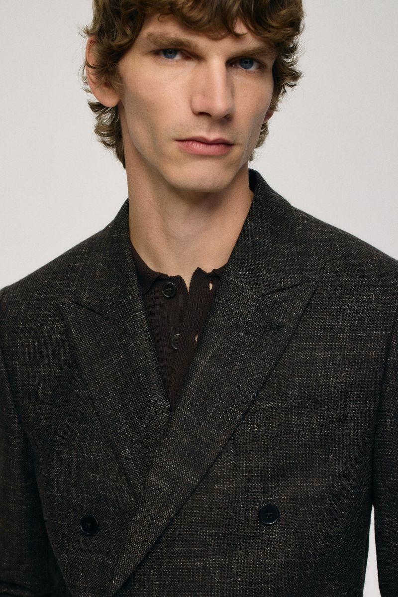 Close-up of a light-skinned adult male with curly brown hair wearing a textured double-breasted gray suit jacket over a brown polo shirt.