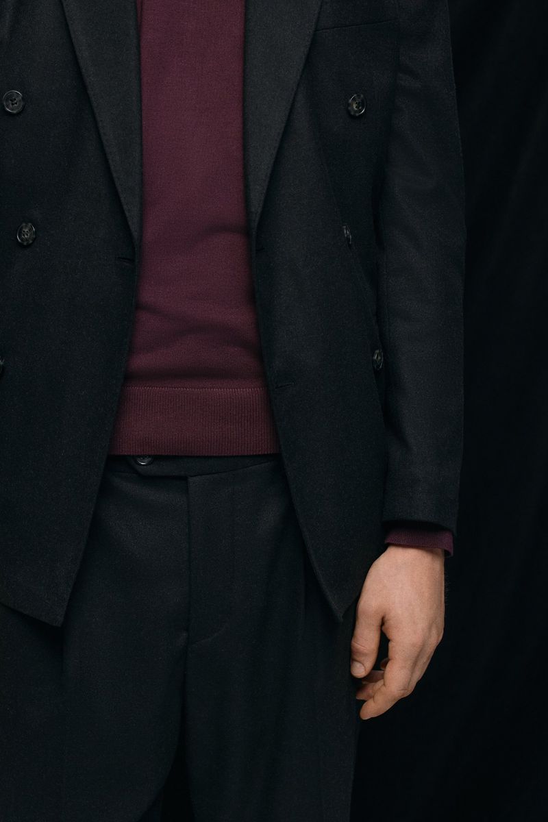 Man in a double-breasted black suit with a burgundy sweater stands against a black backdrop in a fashion style photograph.